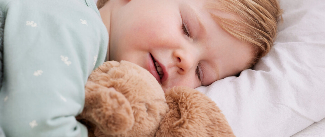 A young child sleeps peacefully on a white pillow, cuddling a brown plush toy.