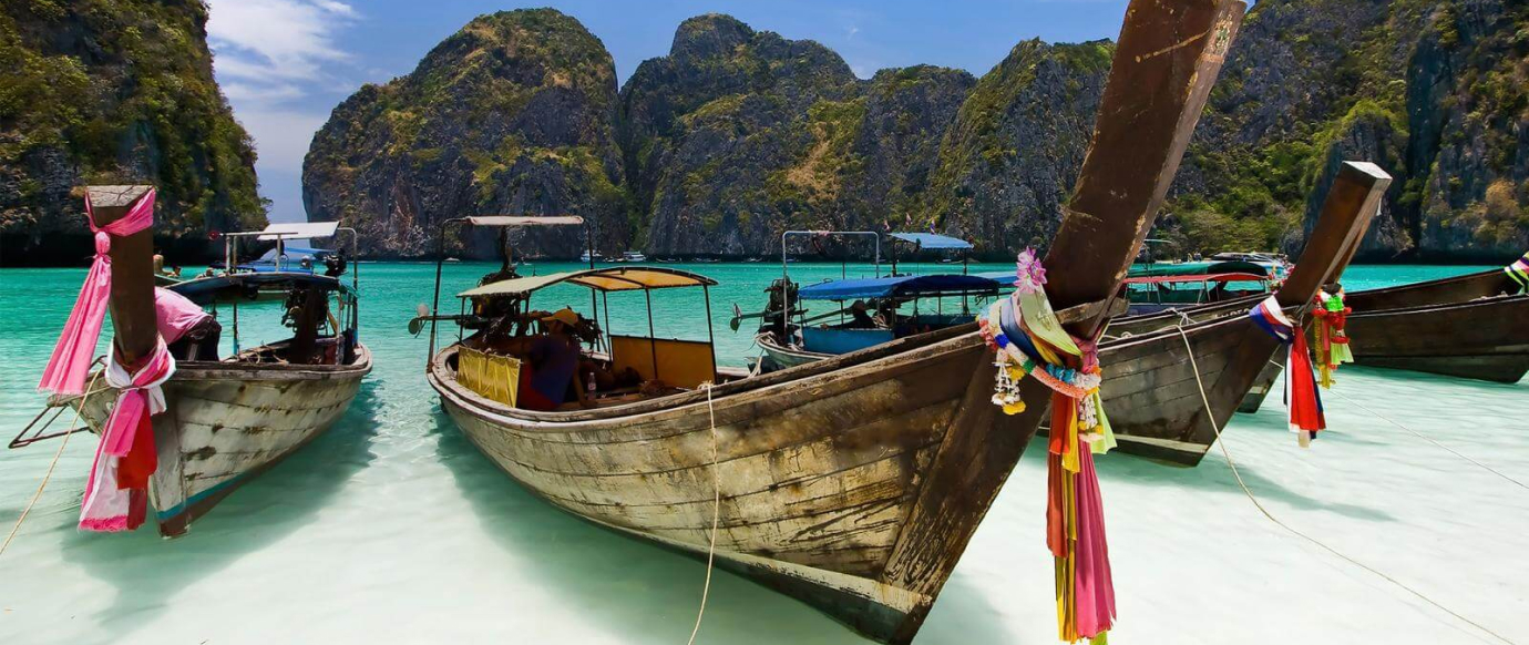 Three wooden boats are docked near a shoreline with turquoise water. Rocky hills covered in greenery form the backdrop. Brightly colored fabric is tied to the bows of the boats.