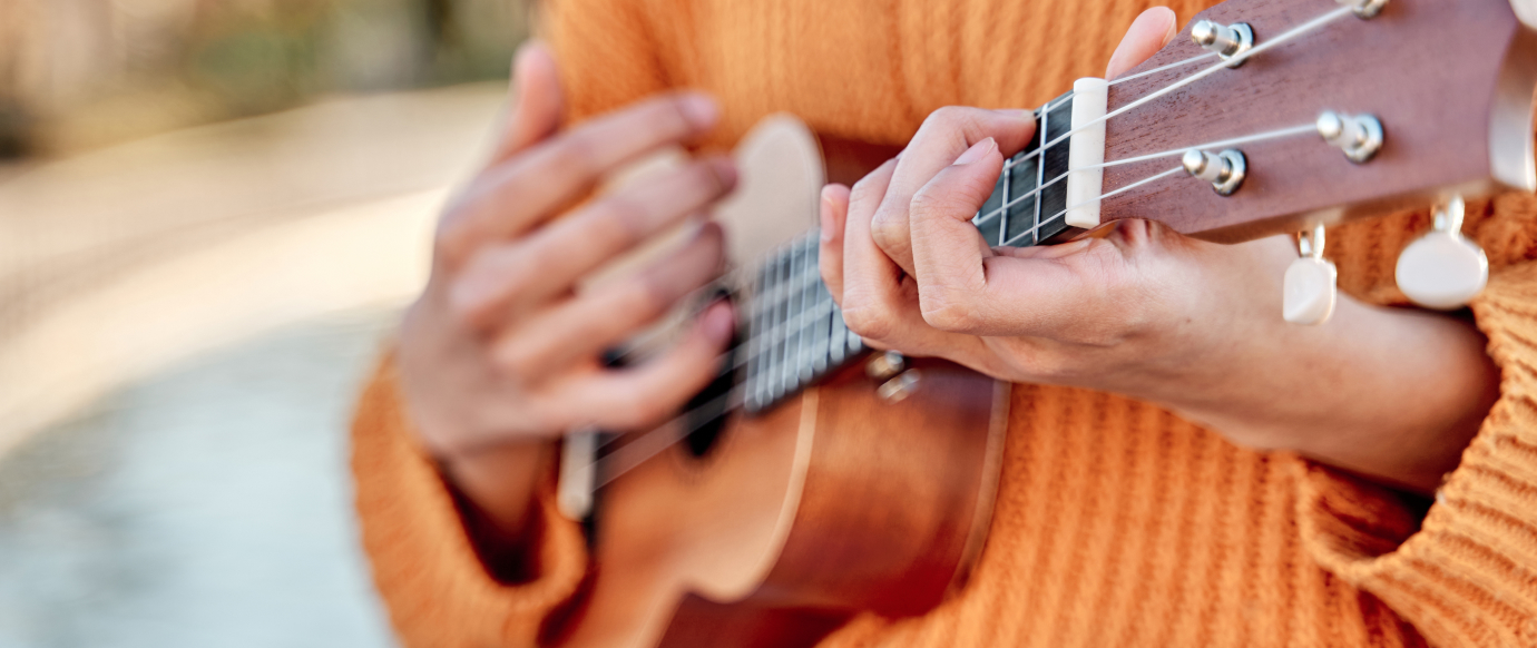 Close-up of hands playing a small stringed instrument, possibly a ukulele, with a background of blurred outdoor scenery and a person wearing an orange sweater.