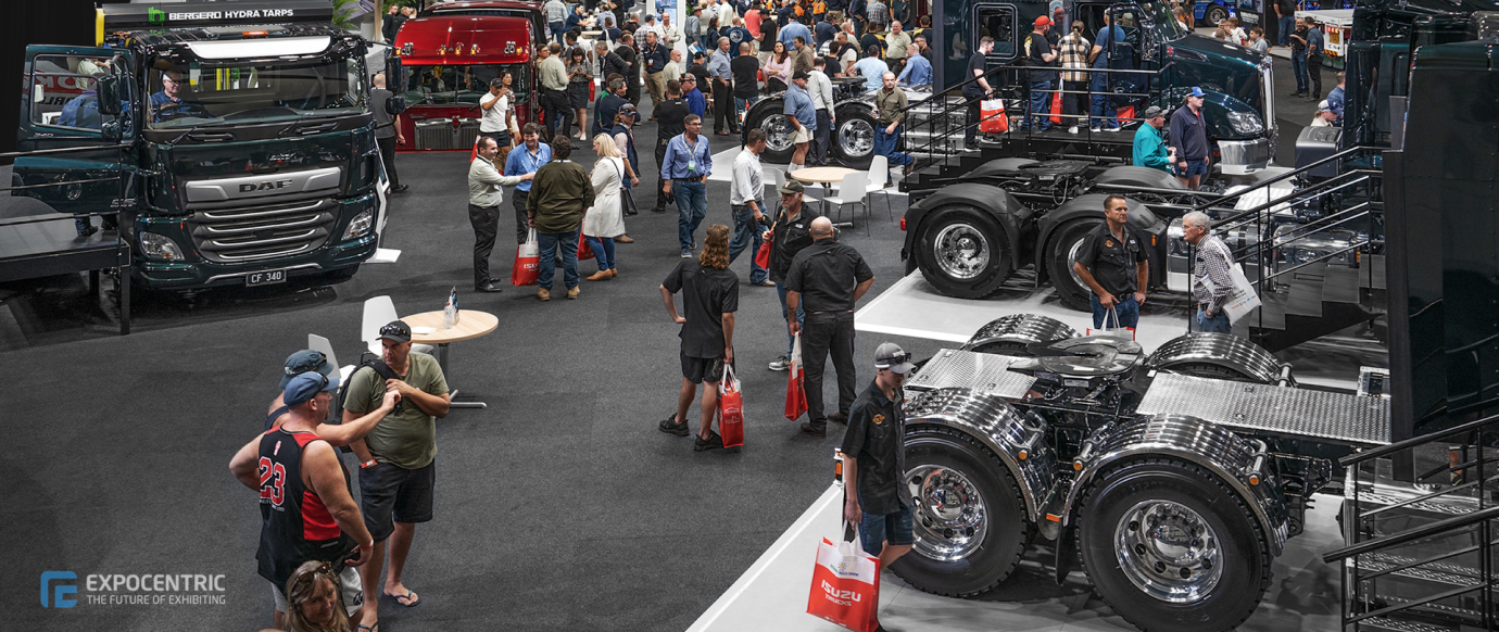 A busy expo floor displaying several trucks and truck parts with visitors interacting and examining the exhibits. The Excentric logo is visible in the bottom left corner.